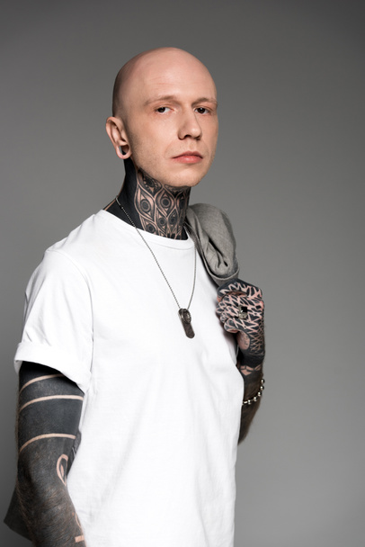 hombre calvo con tatuajes sosteniendo chaqueta de traje en el hombro y mirando a la cámara aislada en gris
 - Foto, imagen