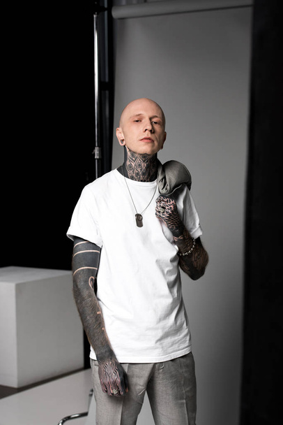 confident bald man with tattoos holding suit jacket on shoulder and looking at camera in studio - Fotografie, Obrázek
