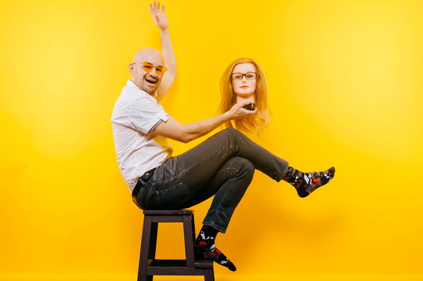 bald man holding plastic mannequin head while sitting on wooden chair on yellow backdrop - Foto, afbeelding