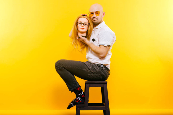bald man holding plastic mannequin head while sitting on wooden chair on yellow backdrop - Фото, зображення