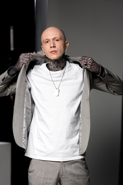 handsome stylish tattooed man wearing suit jacket and looking at camera in studio - Photo, Image