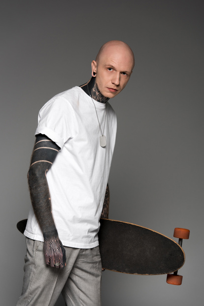 bald tattooed man in white t-shirt holding skateboard and looking at camera isolated on grey - Fotó, kép