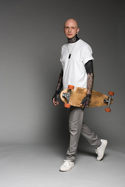 handsome tattooed man in white t-shirt walking with longboard and looking at camera on grey - Foto, imagen