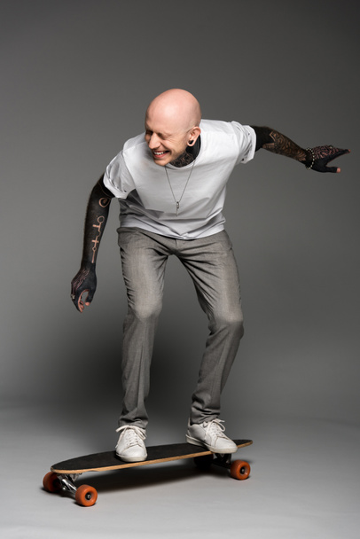 cheerful tattooed man balancing on skateboard and laughing on grey - Фото, зображення
