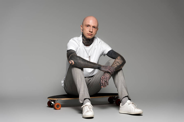 bald tattooed man sitting on skateboard and looking at camera on grey - Zdjęcie, obraz