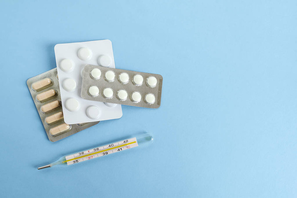Three blisters with pills and capsules and a mercury thermometer on a blue background. To get sick, take pills or undergo treatment. Concept of health care and pharmaceuticals. Flat lay, view from above, copy space. - Фото, изображение