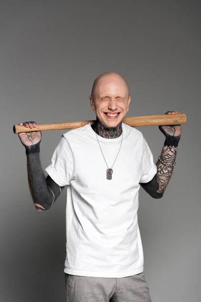 cheerful tattooed man with closed eyes standing with baseball bat on shoulders and laughing on grey  - 写真・画像