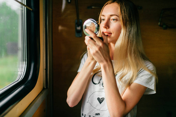 young woman with long hair applying lipstick while travelling by train alone - Фото, зображення