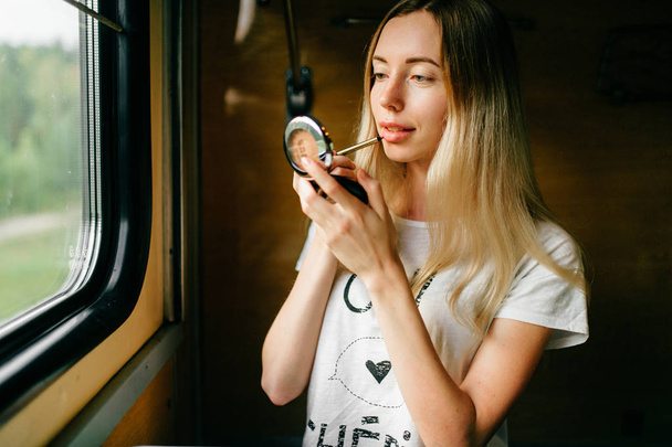 young woman with long hair applying lipstick while travelling by train alone - Foto, imagen