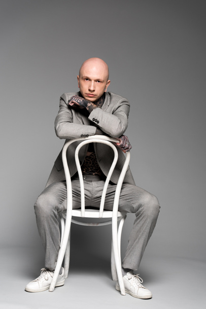 full length view of handsome bald man with tattoos sitting on white chair and looking at camera on grey - Fotografie, Obrázek