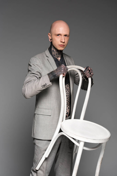 fiducioso uomo pelato tatuato in tuta con sedia bianca e guardando la fotocamera isolata sul grigio
 - Foto, immagini