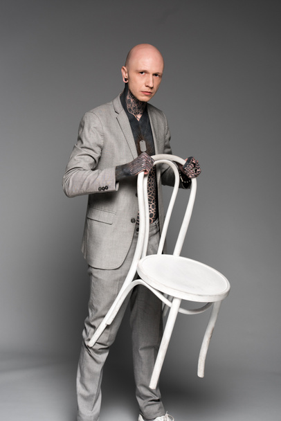 bel homme tatoué sérieux tenant une chaise blanche et regardant la caméra sur gris
 - Photo, image