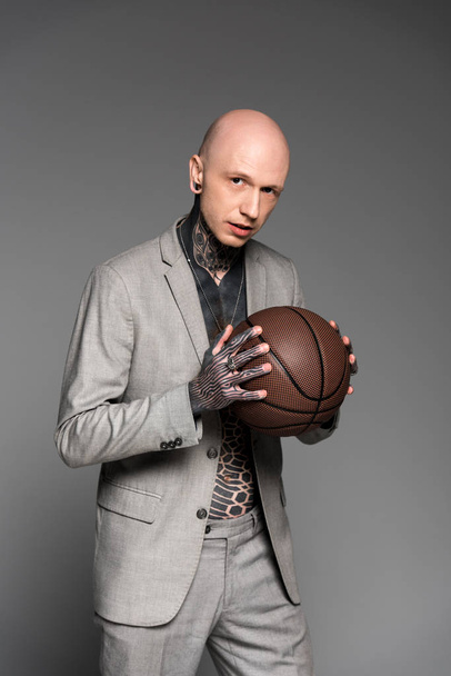 handsome bald tattooed man in suit holding basketball ball and looking at camera isolated on grey  - Foto, immagini