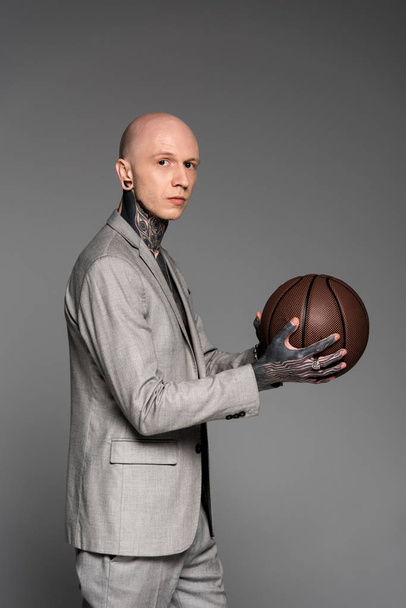 Widok z boku łysy tatuażem człowieka w garniturze, trzymając piłkę do koszykówki i patrząc na kamery na szarym tle - Zdjęcie, obraz