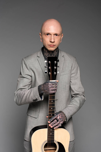 knappe kale tattooed man in pak akoestische gitaar houden en kijken naar camera geïsoleerd op grijs - Foto, afbeelding