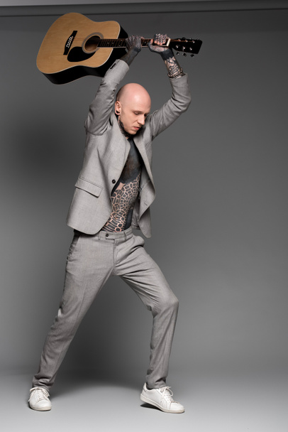full length view of aggressive bald tattooed man in suit hitting acoustic guitar on grey   - Foto, Imagen