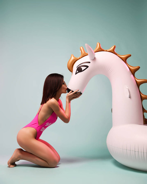 Young fashion woman kissing big inflatable unicorn pegasus float mattress in sexy pink bikini for swimming pool - Фото, зображення