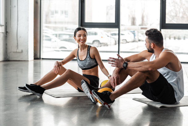 yoga paspaslar üzerine oturan ve spor salonunda konuşurken spor giyim atletik genç çift - Fotoğraf, Görsel