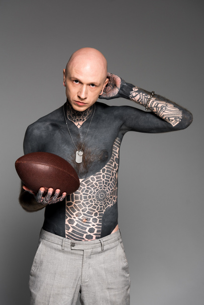 bald shirtless tattooed man holding rugby ball and looking at camera isolated on grey - Foto, immagini