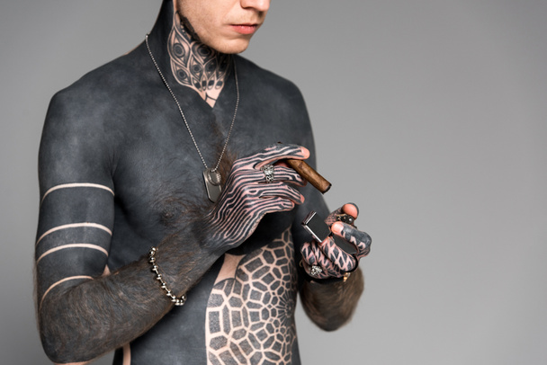 cropped shot of bare-chested man with tattoos holding cigar and lighter isolated on grey - Фото, изображение