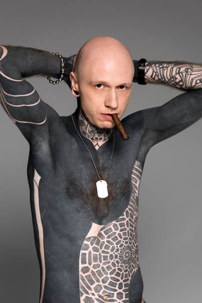 confident tattooed bare-chested man with hands behind head smoking cigar and looking at camera isolated on grey - Zdjęcie, obraz