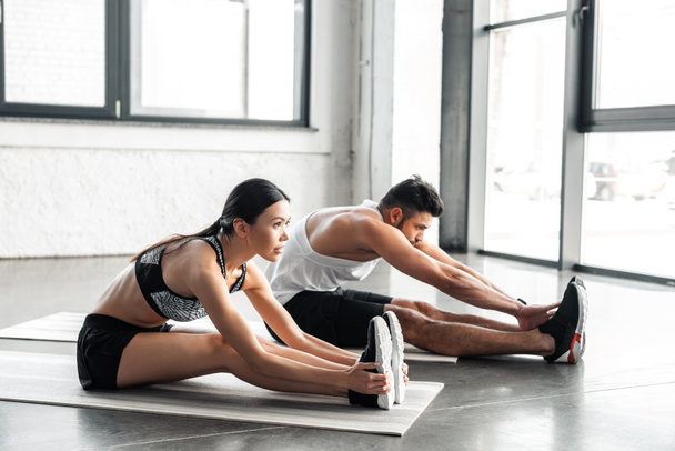vista lateral de pareja joven y deportiva estirándose y ejercitándose sobre colchonetas de yoga en el gimnasio
   - Foto, imagen