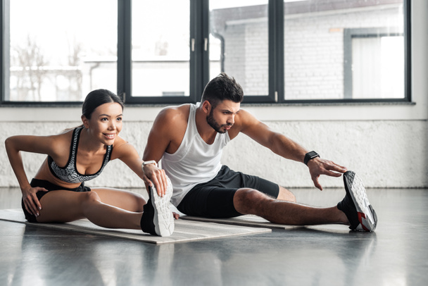 athletic young man and woman stretching legs on yoga mats in gym - Photo, Image