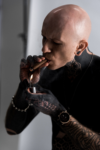 calvo sin camisa hombre tatuado sosteniendo más ligero y fumar cigarro en gris
 - Foto, Imagen