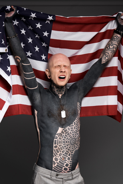 emotional tattooed man with closed eyes holding us flag isolated on grey - 写真・画像