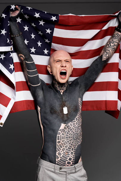 emocionado sin camisa hombre de pecho desnudo con tatuajes sosteniendo bandera americana y gritando aislado en gris
  - Foto, Imagen
