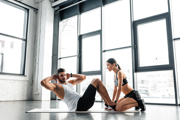 side view of young sportswoman holding legs of athletic man doing abs exercise in gym - Photo, Image