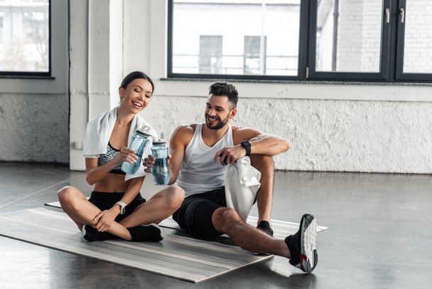улыбающаяся молодая пара в спортивной одежде, сидящая на ковриках для йоги и держащая бутылки с водой после тренировки в спортзале
 - Фото, изображение