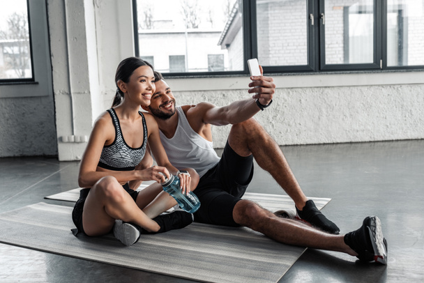 Happy νεαρό ζευγάρι σε αθλητικά ενδύματα λαμβάνοντας αυτοπορτρέτα με smartphone ενώ στηρίζεται σε χαλάκια γιόγκα για μετά την προπόνηση στο γυμναστήριο  - Φωτογραφία, εικόνα