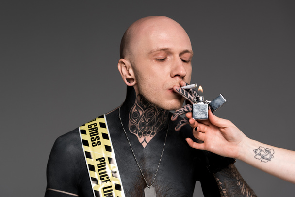 partial view of someone holding lighter and tattooed man with cross line around body smoking roll-up cigarette isolated on grey - Foto, Bild