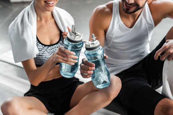 cropped shot of smiling young couple in sportswear holding bottles of water while sitting and resting in gym - Foto, Imagem