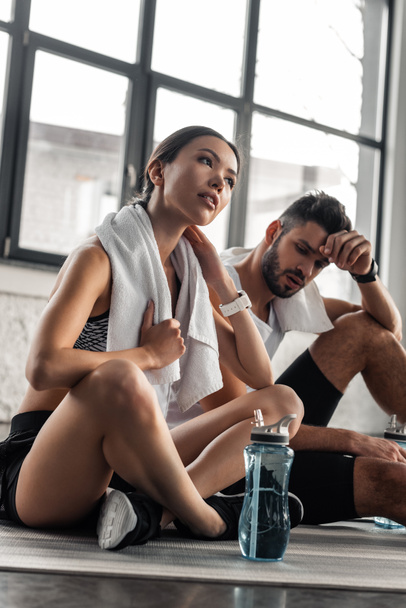 low angle view of tired sporty young couple with towels and bottles of water resting on yoga mats in gym - Фото, зображення