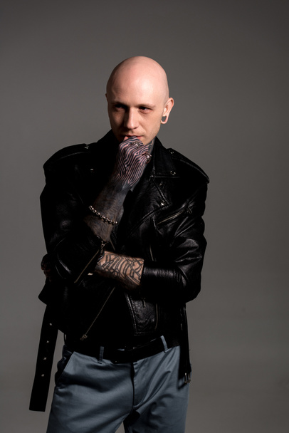 calvo hombre tatuado en chaqueta de cuero de pie con la mano en la barbilla y mirando hacia otro lado aislado en gris
 - Foto, Imagen