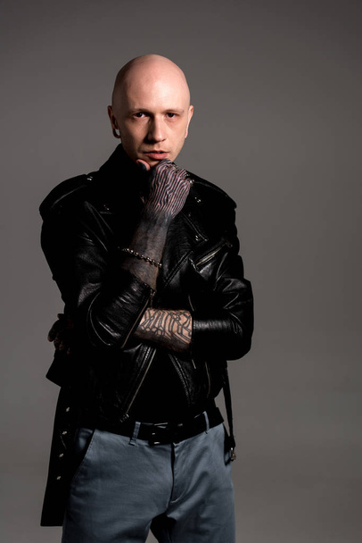 stylish tattooed man in leather jacket standing with hand on chin and looking at camera isolated on grey - Photo, Image