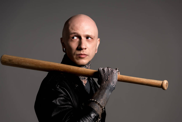 bald tattooed man in leather jacket holding baseball bat and looking away isolated on grey - Foto, Imagen