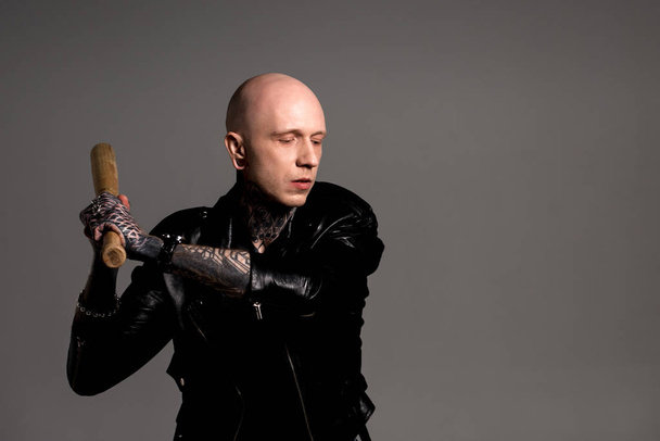 aggressive tattooed man in leather jacket hitting with baseball bat isolated on grey  - Fotografie, Obrázek
