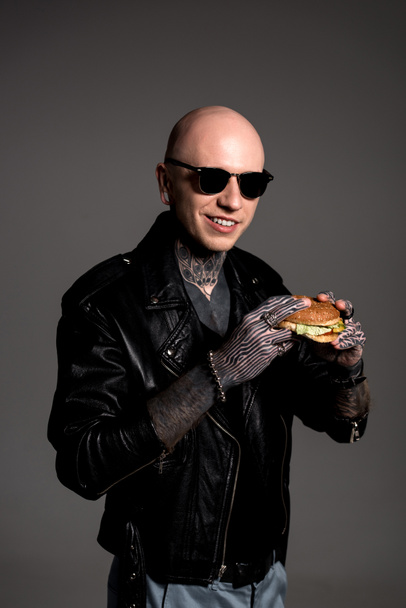 uomo tatuato in giacca di pelle e occhiali da sole che tiene gustoso hamburger e sorride alla telecamera isolata su grigio
  - Foto, immagini