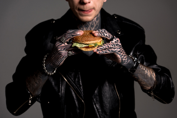 cropped shot of hungry tattooed man in leather jacket holding burger isolated on grey - Photo, Image