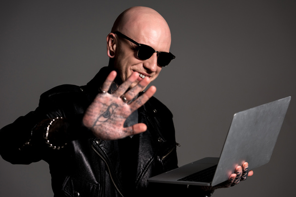 glimlachend kale man in leren jas en zonnebril holding laptop en weergegeven: palm met tatoeages geïsoleerd op grijs - Foto, afbeelding