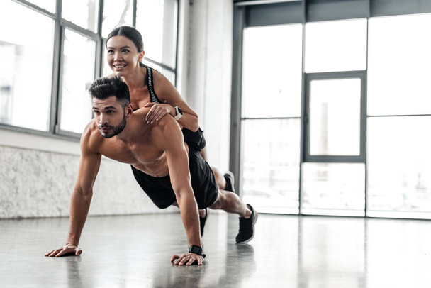 muskulöser Sportler mit nacktem Oberkörper beim Liegestütz mit lächelndem sportlichem Mädchen auf dem Rücken im Fitnessstudio  - Foto, Bild