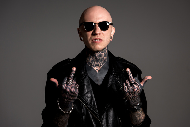 uomo tatuato aggressivo in giacca di pelle e occhiali da sole con dita medie isolate sul grigio
 - Foto, immagini