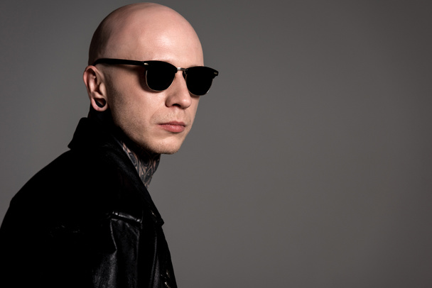 stylish tattooed guy in leather jacket and sunglasses posing isolated on grey - Foto, afbeelding
