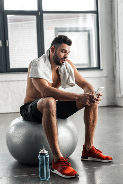 young muscular bare-chested sportsman sitting on fit ball and using smartphone in gym - Foto, afbeelding