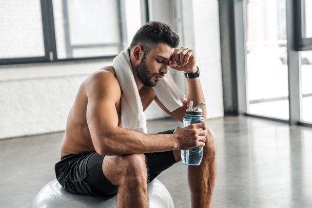 tired muscular sportsman with towel and bottle of water sitting on fitness ball in gym - Photo, Image
