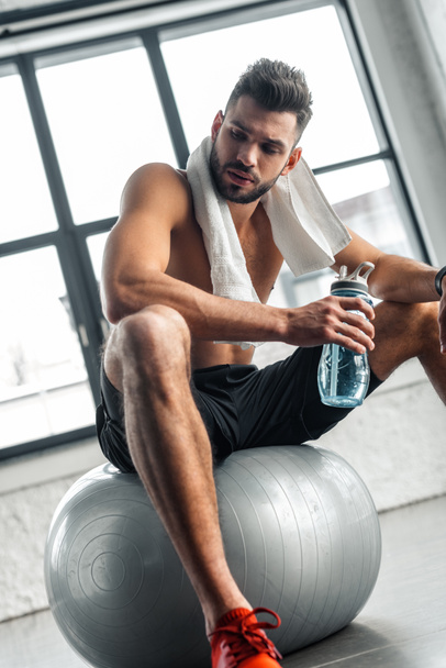 young bare-chested sportsman with towel and bottle of water sitting on fit ball in gym - Photo, Image