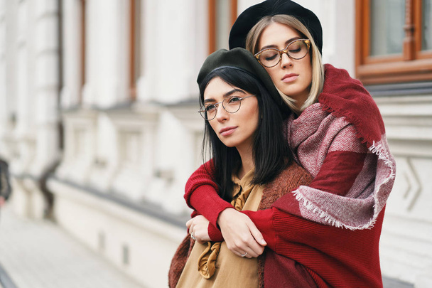 Two female friends outdoors portrait. Girls in casual warm outfits, glasses and berets sitting and hugging on urban background, copy space. City lifestyle, friendship and sisterhood concept.  - Foto, immagini